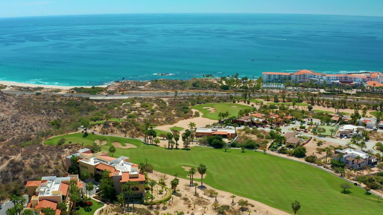 Opening aerial frame over Marquis Los Cabos, Mexico, with golf greens and homes in the foreground, a coastal road near the beach, blue ocean beyond, and no visible people.