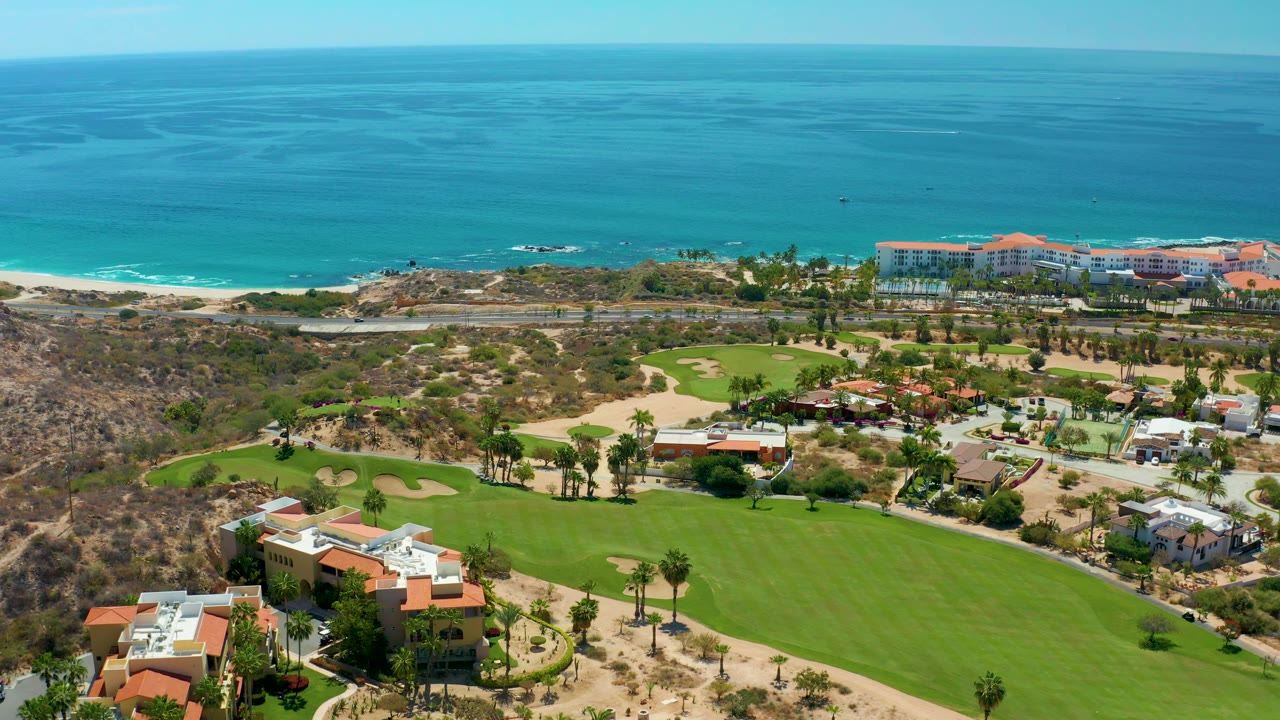 One second into the clip, the aerial view holds on Marquis Los Cabos with manicured golf greens below, dry terrain and a road in the middle distance, ocean surf behind, and no visible people.