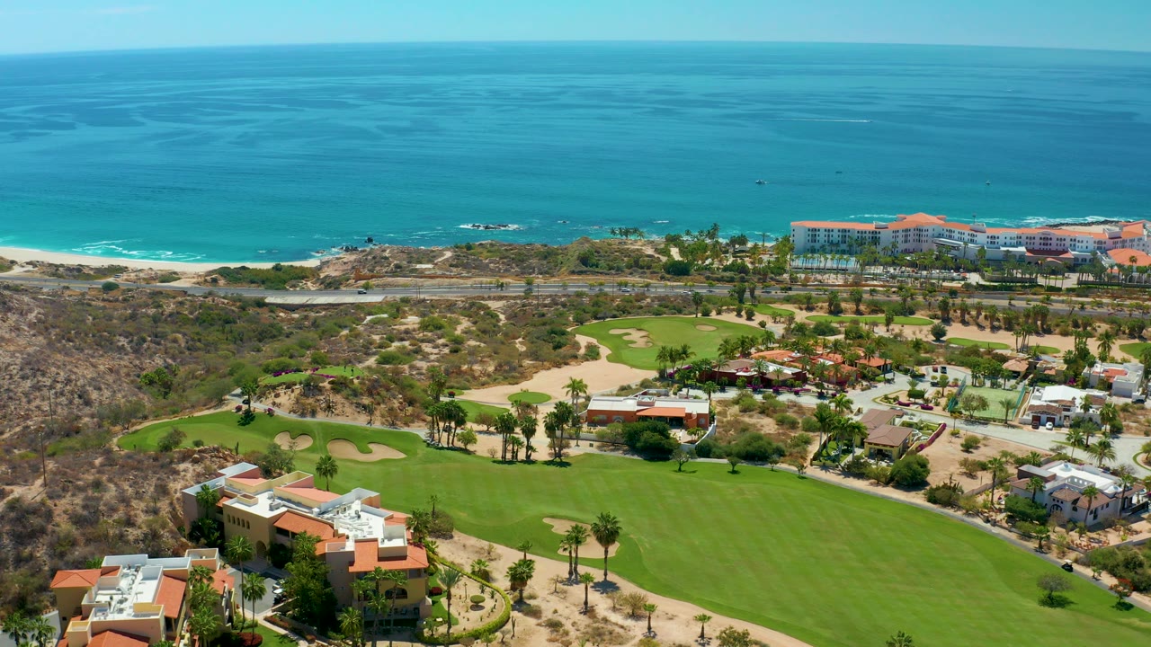 At 0:02, Marquis Los Cabos appears from above with villas and palms around the golf course, a road running parallel to the beach, small vehicles, blue water, and no visible people.