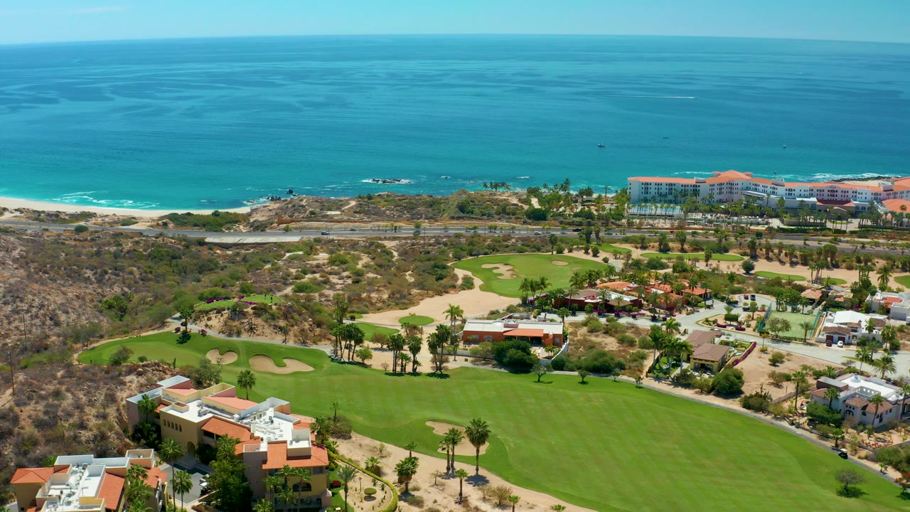 Aerial view of green fairways, sandy terrain, buildings, beach, and ocean horizon.