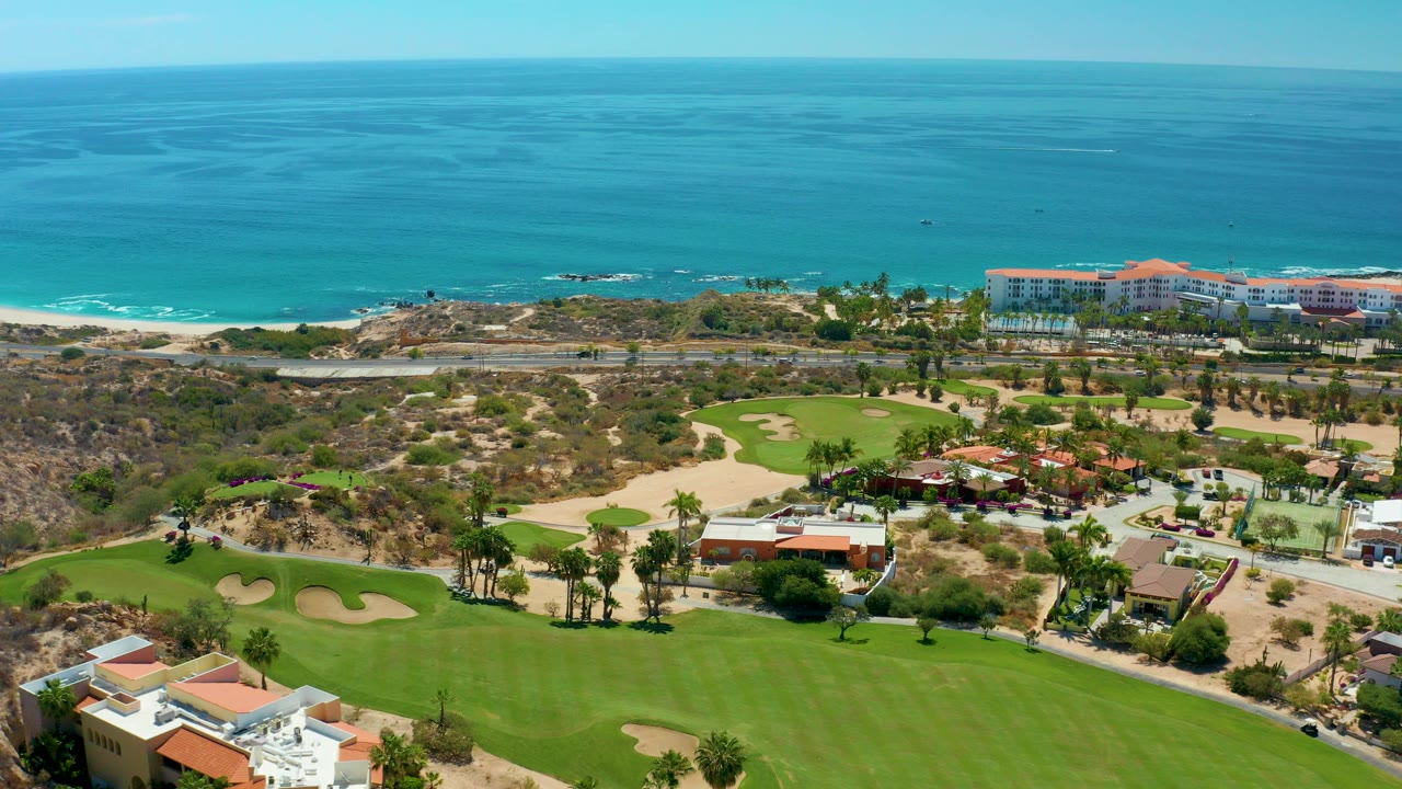 Opening aerial frame at Marquis Los Cabos, Mexico, with green fairways, sand bunkers, palm trees, resort-area buildings, beach surf, and blue ocean, with no visible people.