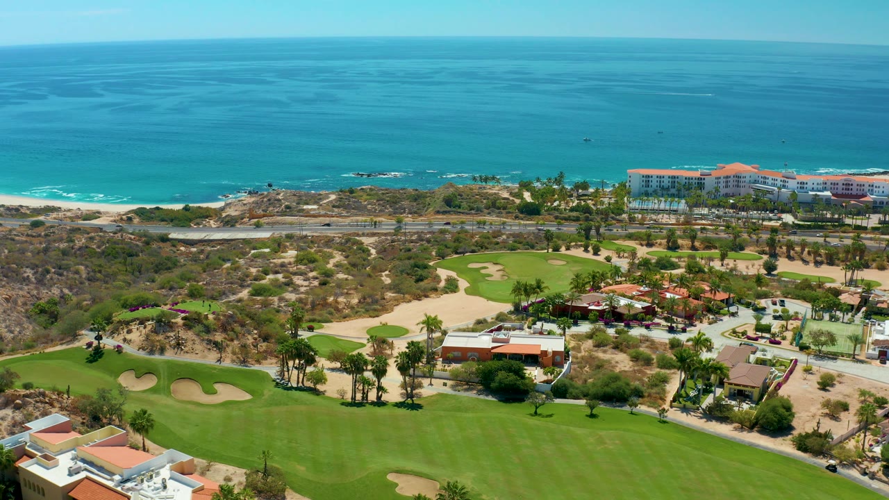 Aerial frame at Marquis Los Cabos, Mexico, with a broad fairway in the foreground, clustered red-roof buildings and palms in the middle distance, and turquoise ocean behind, with no visible people.