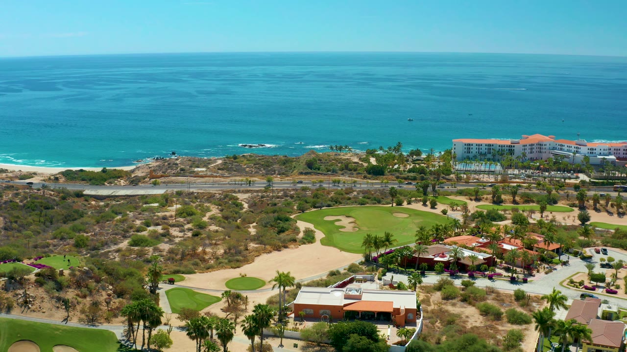 Wide aerial frame at Marquis Los Cabos, Mexico, with the blue ocean across the top, the shoreline road below it, fairways and bunkers in front, and no visible people.