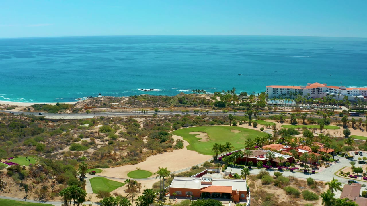 At Marquis Los Cabos, Mexico, this aerial frame shows a golf course foreground, a road paralleling the shore, low surf on the beach, open ocean, and no visible people.