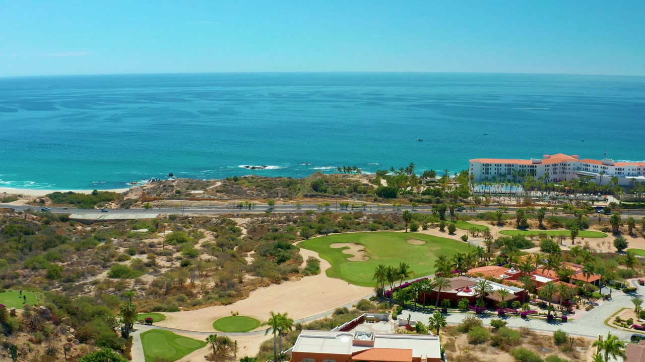 Aerial view at Marquis Los Cabos, Mexico, with coastal buildings near the shoreline, a road with small vehicles, a green golf course below, and no visible people.
