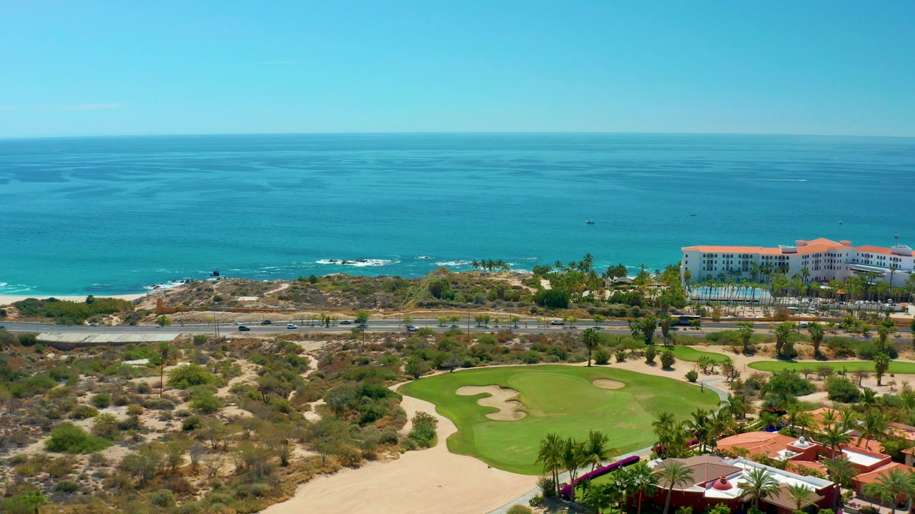 Opening aerial frame at Marquis Los Cabos, Mexico showing golf greens and sand traps below, a coastal road, red-roof resort buildings, blue ocean, clear sky, and no visible people.