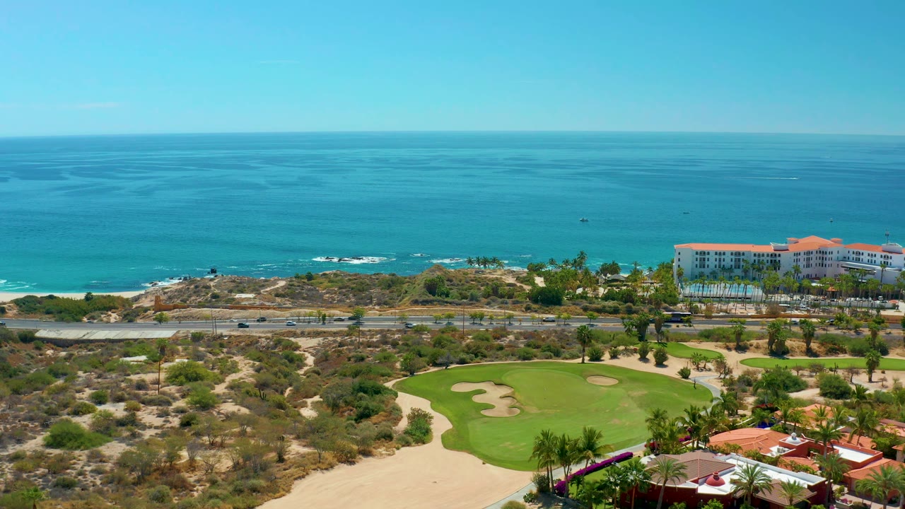Wide aerial coastline with golf green, road, resort buildings, and calm ocean.
