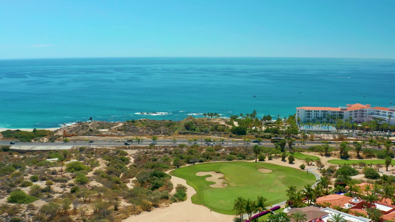 Aerial view at Marquis Los Cabos, Mexico with a curved golf green and sand traps in the foreground, cars on the coastal road, red-roof resort buildings to the right, ocean, and no visible people.