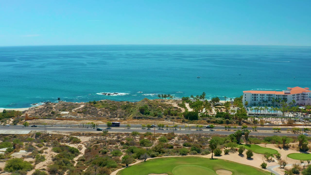 Aerial frame at 0:01 over Marquis Los Cabos BCS Mexico with the highway lower in frame, cars spaced along the lanes, ocean behind the coastal scrub, and no visible people.