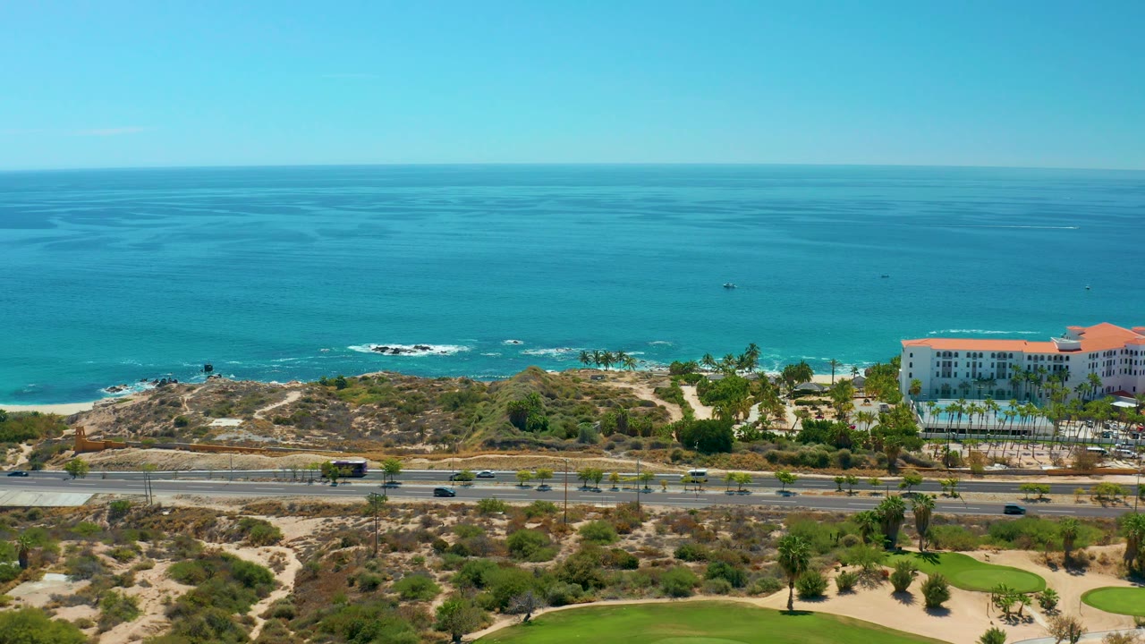 Aerial frame at 0:02 from Marquis Los Cabos BCS Mexico showing more open water across the background, light traffic on the coastal highway, green fairway areas in front, and no visible people.