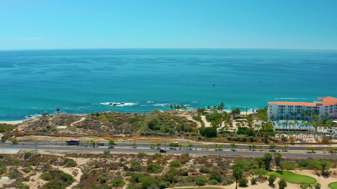 Oceanfront highway scene with cars, scrub-covered coast, palms, and red-roofed building.