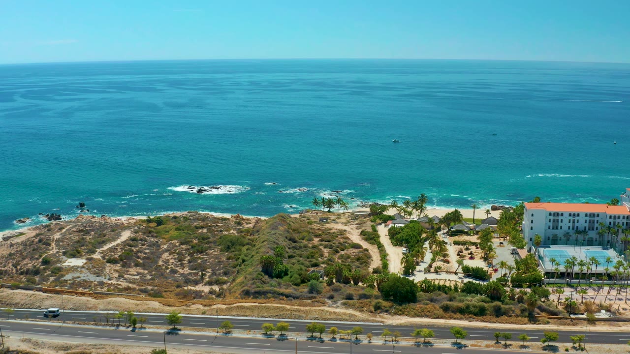 Aerial frame at Marquis Los Cabos, Mexico with the ocean horizon high in frame, surf on rocks, sandy shoreline, scrub-covered ground, a road below, and a white building and pool entering at right with no visible people.