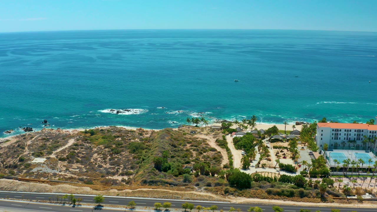 Aerial frame at Marquis Los Cabos, Mexico showing calm blue water, small wave lines, a pale beach, dry coastal brush, a road along the bottom edge, and more of the white building and pool visible at right with no visible people.