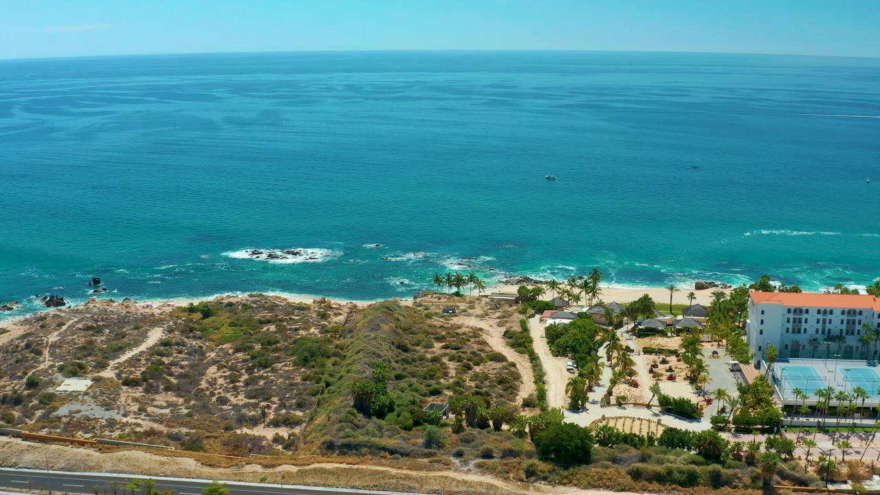 Aerial frame at Marquis Los Cabos, Mexico with turquoise water across the upper frame, surf reaching the beach, branching sandy paths through dry vegetation, a road in front, and the pool area near the right edge with no visible people.