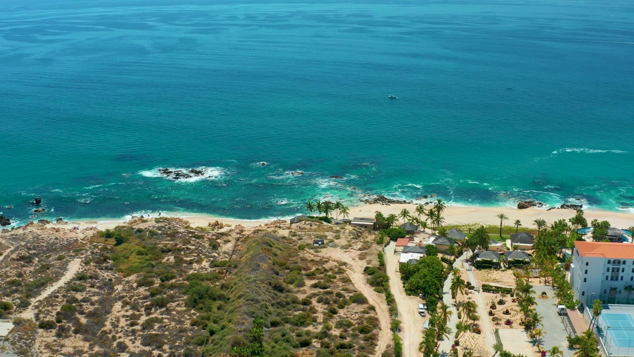 Aerial frame from Marquis Los Cabos, Mexico showing turquoise water, white surf over dark rocks, sandy beach, dry scrub hills, palms, and coastal buildings with no visible people.