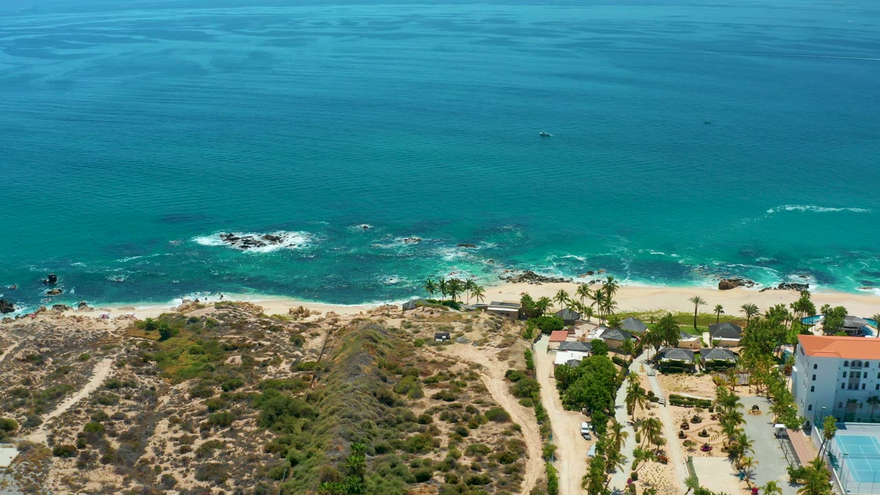 Aerial view at Marquis Los Cabos, Mexico with foamy waves spread across nearshore rocks, a narrow sandy beach, dry hillside paths, palm trees, and a white coastal building with no visible people.