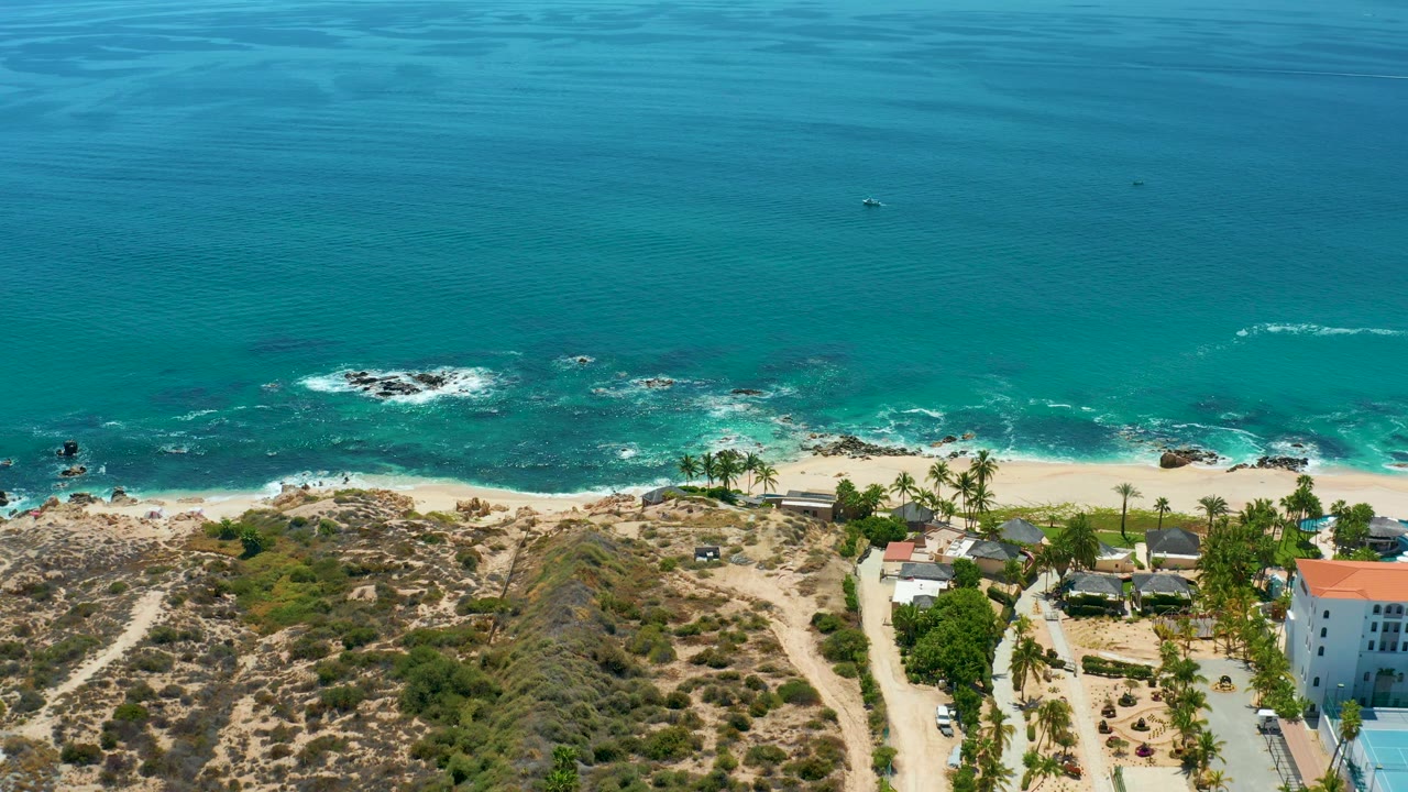 High aerial frame from Marquis Los Cabos, Mexico showing blue-green ocean, white water over rocks, a sandy shore, sparse dry vegetation, palm clusters, and coastal buildings with no visible people.