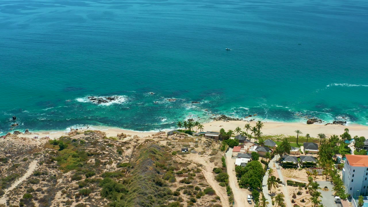 Aerial shoreline with turquoise water, breaking waves, dry hills, and coastal structures.