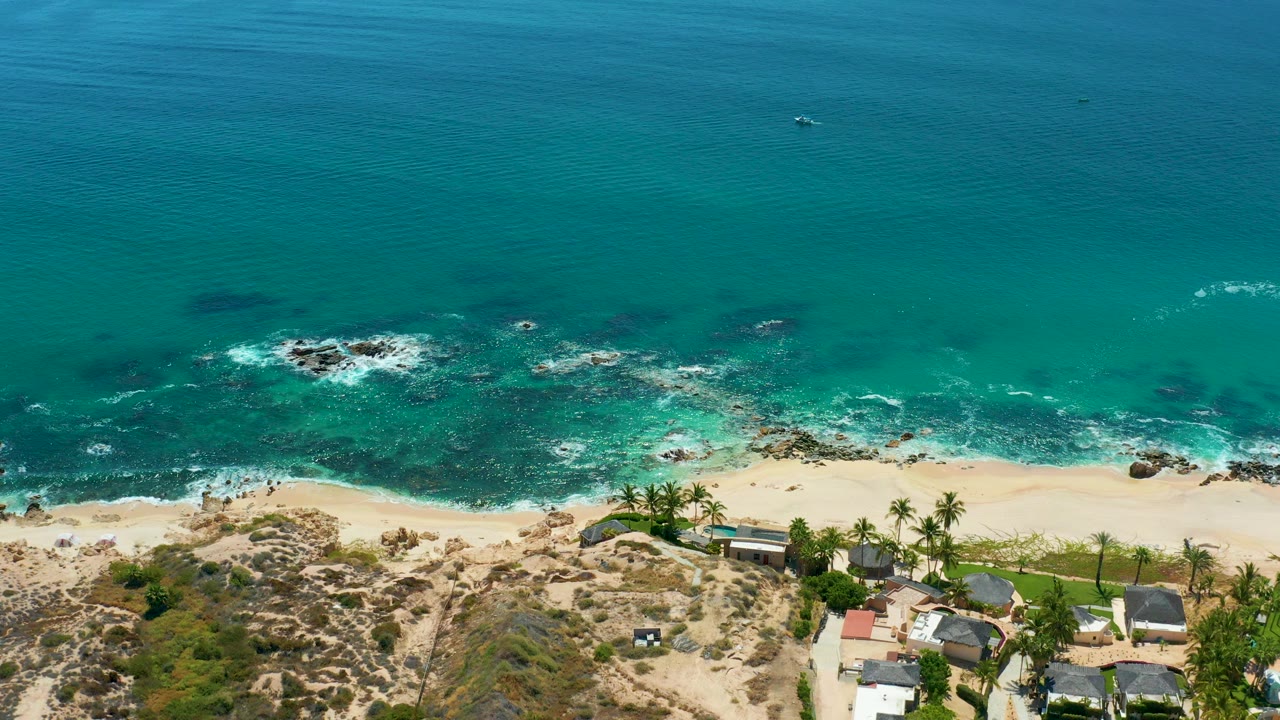 Opening aerial frame over Marquis Los Cabos, Mexico, with turquoise water above dark rocks, white surf at the sand, coastal buildings and palms at the lower edge, and no visible people.