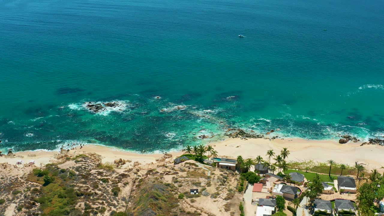Aerial frame at Marquis Los Cabos, Mexico, showing blue-green ocean, foam spreading around offshore rocks, pale sand below, low buildings near palms, and no visible people.