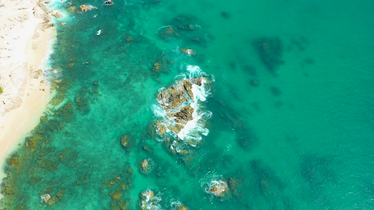 Aerial view of turquoise water, sandy shore, rocks, and white wave foam.