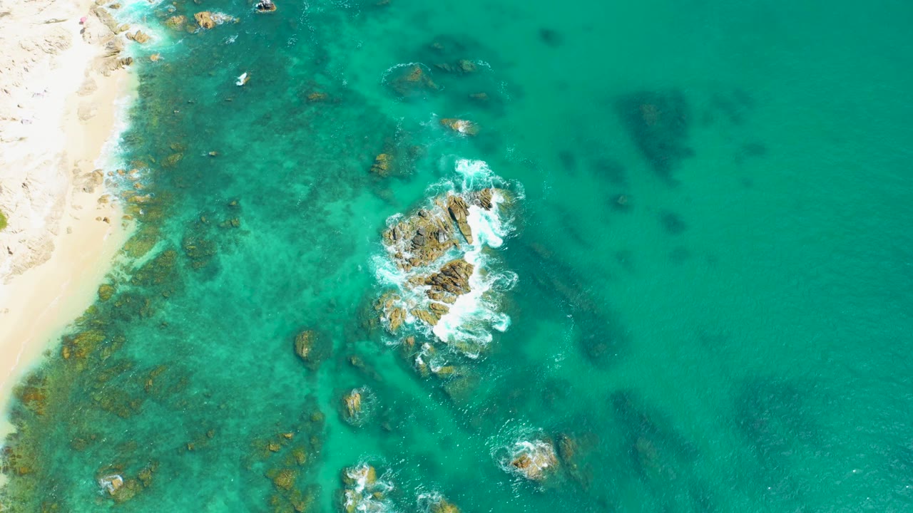 Aerial frame over Marquis Los Cabos, Mexico, showing the beach edge at left, reef-like rock patches under the water, white foam curling around the central rocks, and no visible people.
