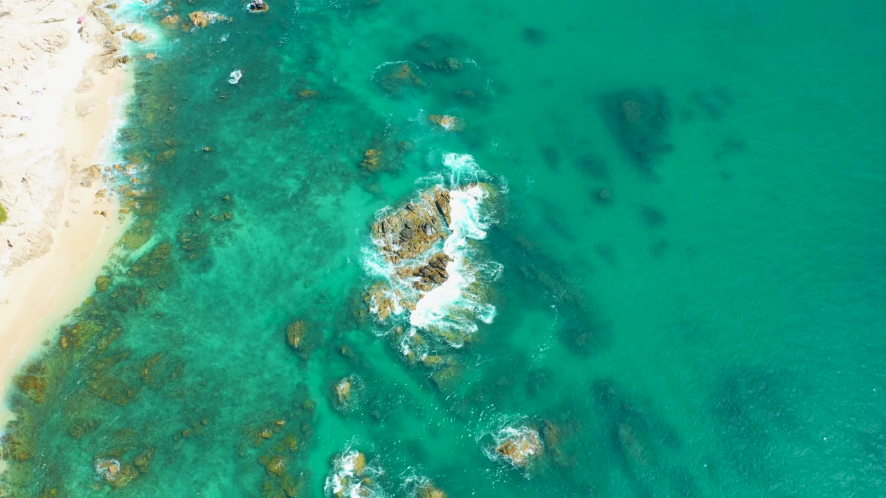 Top-down Marquis Los Cabos coastal view with the sandy strip on the left, open turquoise water to the right, scattered underwater rocks, breaking white surf on the central outcrop, and no visible people.