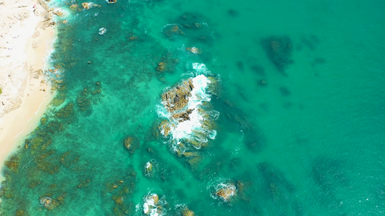 Overhead view at Marquis Los Cabos, Mexico, where a larger patch of white foam spreads across the central rocks, with sandy shore at left, clear shallow water, darker submerged areas, and no visible people.