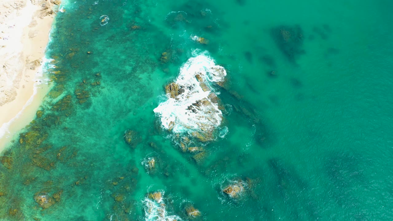 Aerial frame from Marquis Los Cabos, Mexico showing white foam expanding over the main rocks, darker underwater patches around them, sand along the left edge, and no visible people.