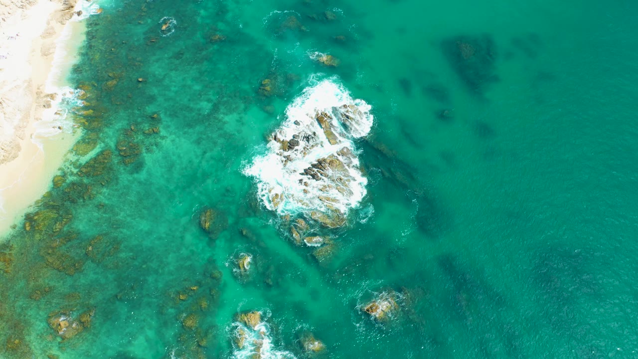 Drone view over Marquis Los Cabos, Mexico with surf wrapping around the central rock cluster, visible reef-like patches in the turquoise shallows, sand at far left, and no visible people.