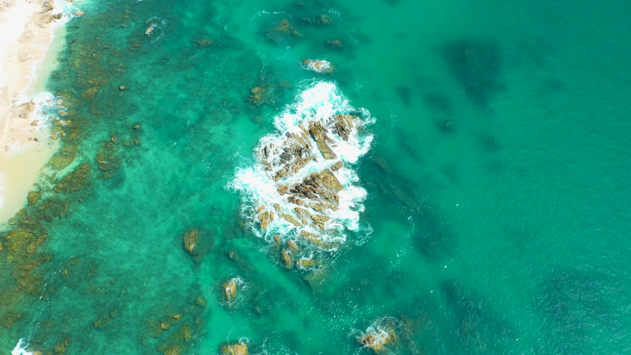 At 0:01, Marquis Los Cabos surf spreads over the central rocks while darker submerged patches sit beneath the clear turquoise water, with no visible people.