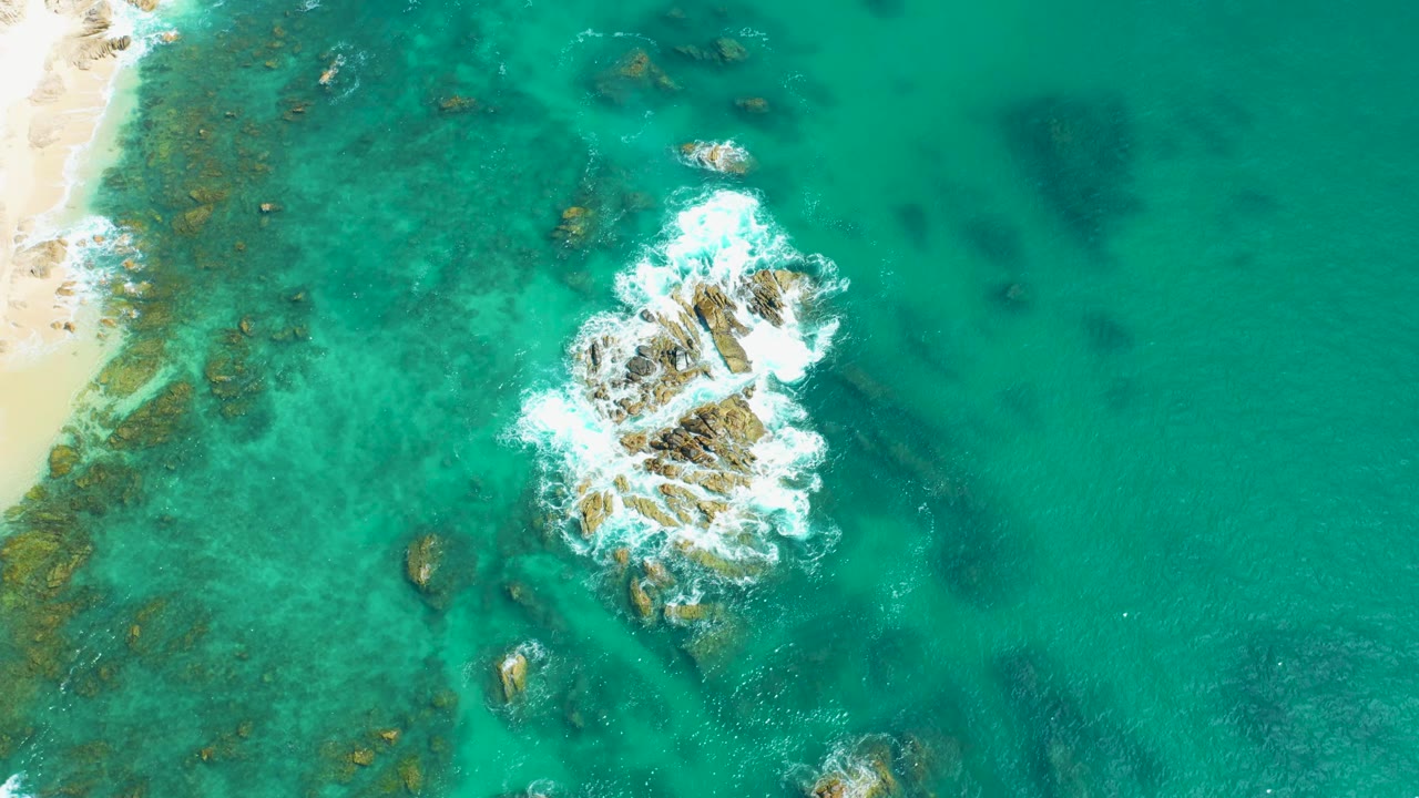 At 0:02, the aerial view holds the rocky outcrop below a bright patch of breaking foam, with pale sand visible at the left edge and no visible people.
