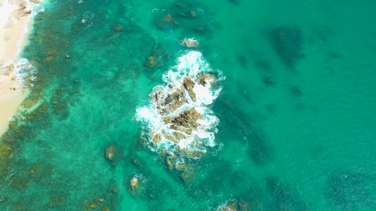 At 0:03, the central rocks sit lower in frame as white water curls around them and the sandy beach remains along the left margin, with no visible people.