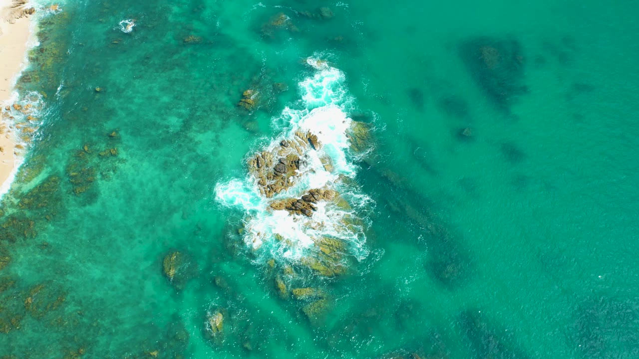 Overhead frame at Marquis Los Cabos, Mexico, showing white surf spreading across a central rock cluster with reef texture and sand visible at the left, no visible people.