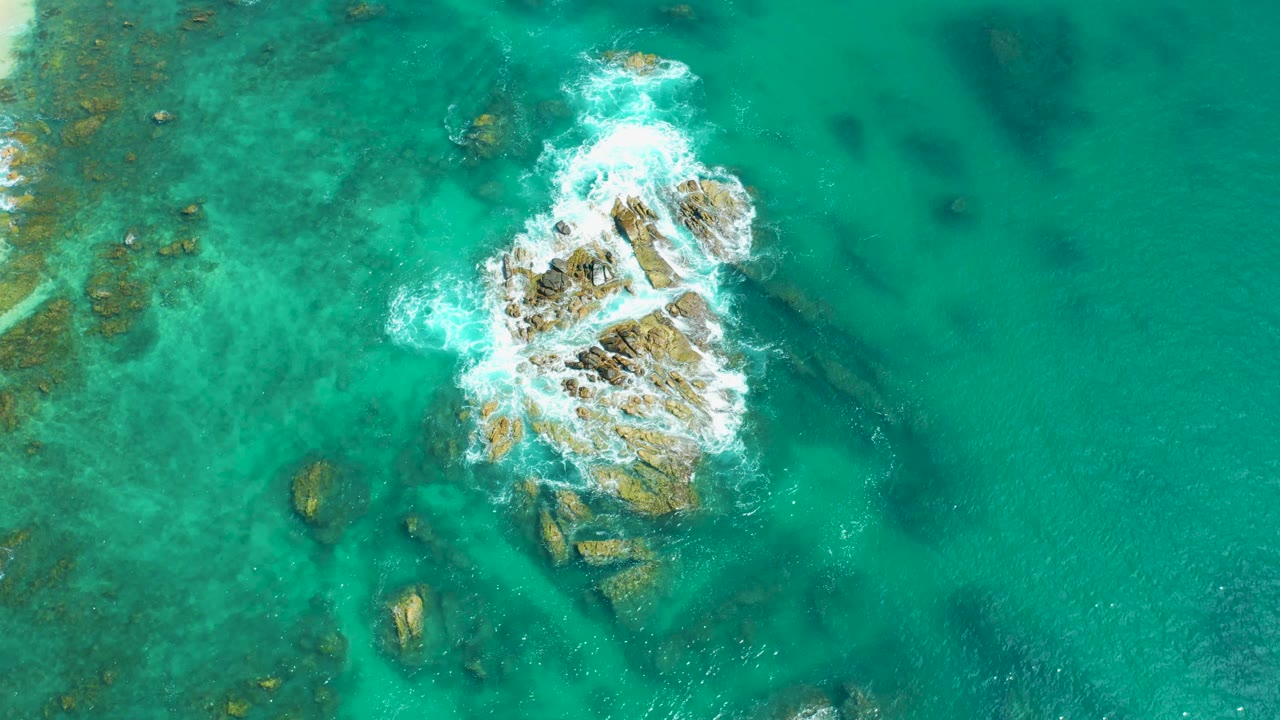 Aerial frame at Marquis Los Cabos, Mexico showing a central rock formation with bright white wave foam across the top and darker underwater patches nearby, with no visible people.