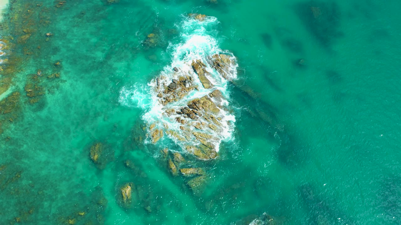 Top-down drone-style view of the Marquis Los Cabos rocky outcrop with foam wrapping around the upper rocks and clear green-blue water revealing seabed texture, with no visible people.