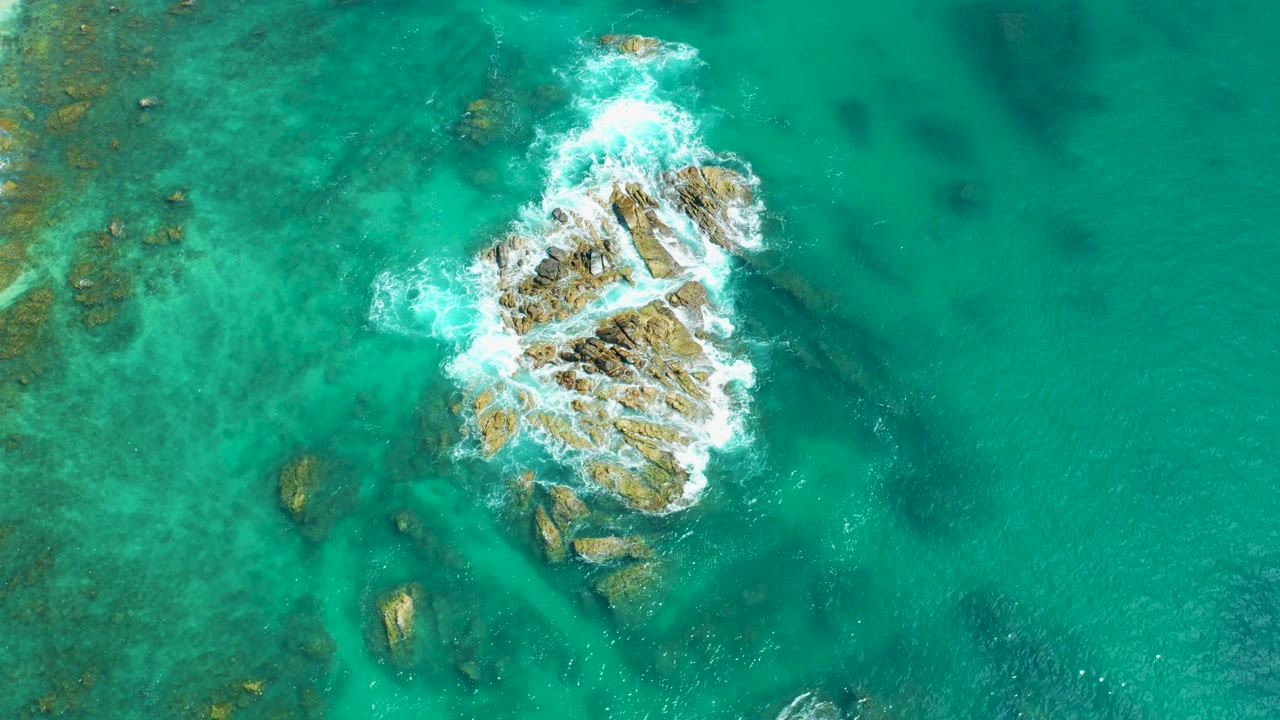 Aerial frame from Marquis Los Cabos, Mexico with tan rocks near center, white water washing over their left and upper edges, and translucent turquoise sea around them, with no visible people.