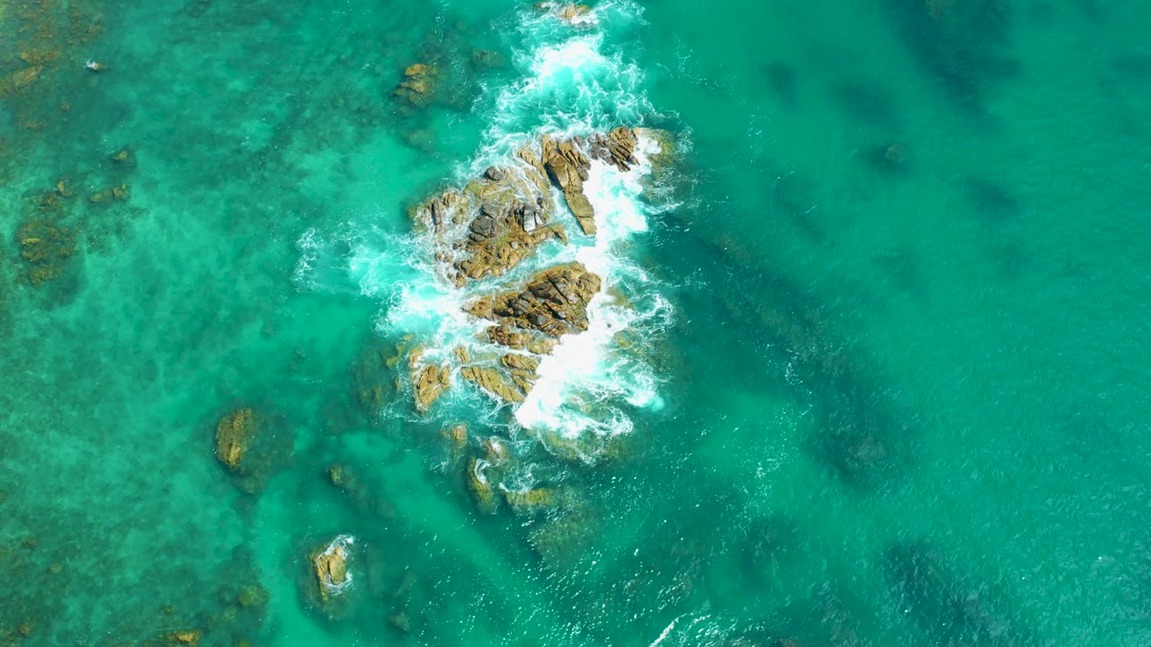 Aerial frame from Marquis Los Cabos, Mexico, with white surf spreading across the upper side of the central rocks and darker underwater patches visible nearby, with no visible people.