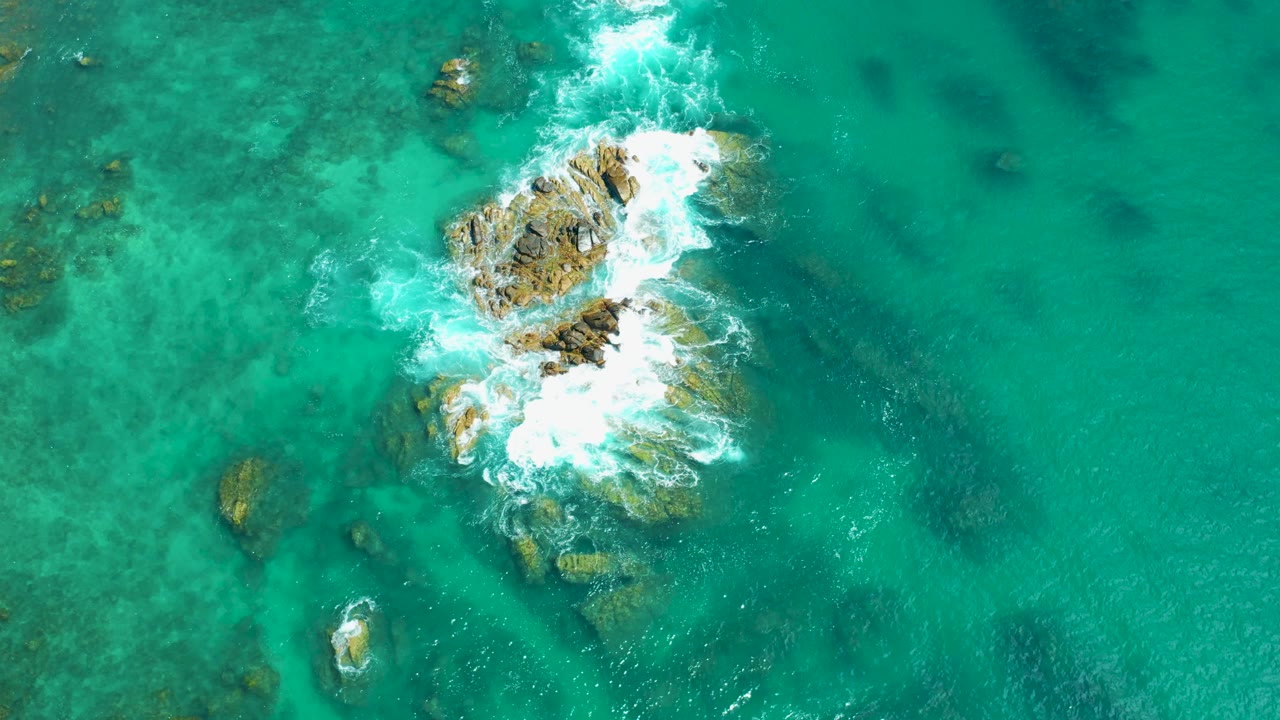 Aerial frame from Marquis Los Cabos, Mexico, showing whitewater washing farther across the central rocks with a smaller isolated rock below, with no visible people.