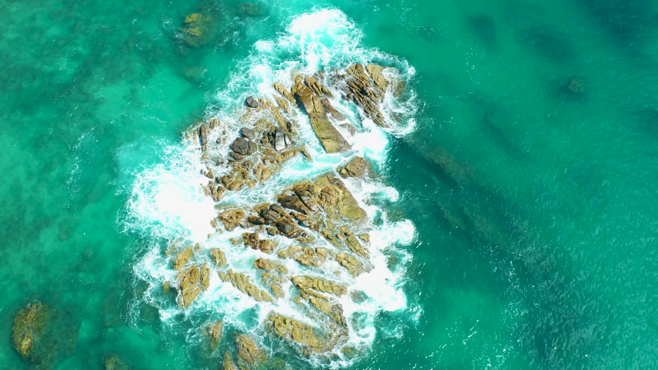 Overhead frame from Marquis Los Cabos, Mexico showing a brown rocky outcrop surrounded by turquoise water and white surf, with no visible people.