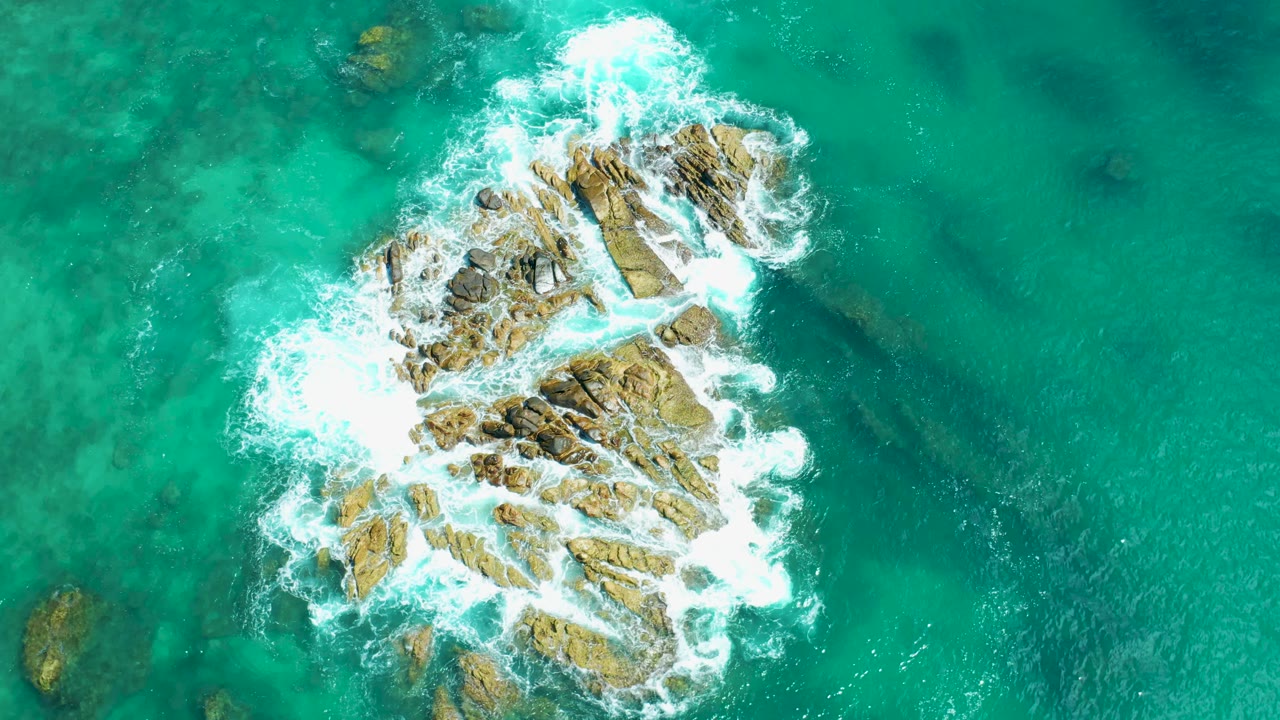 Aerial view at Marquis Los Cabos, Mexico with white water pushing over the upper side of the central rocks and clear green-blue water around them, no visible people.