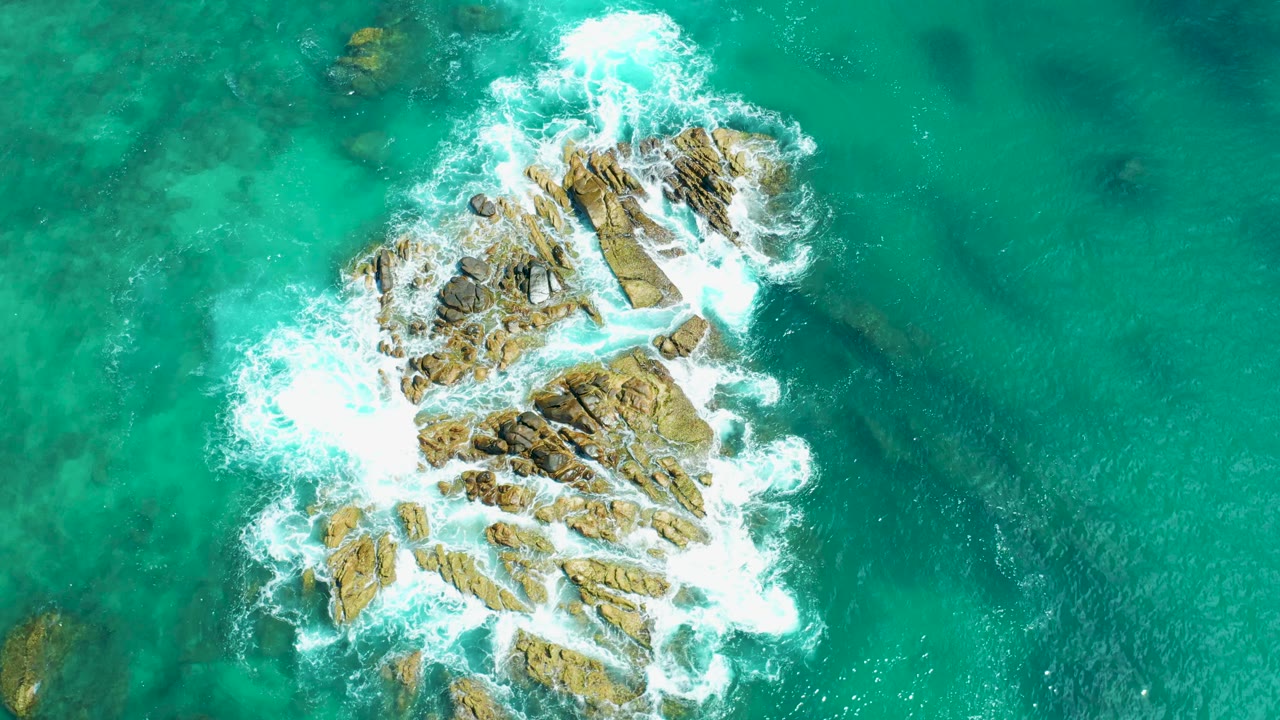 Near-vertical drone frame from Marquis Los Cabos, Mexico showing the rocky outcrop shifted toward the right as surf breaks along its upper and left edges, with no visible people.
