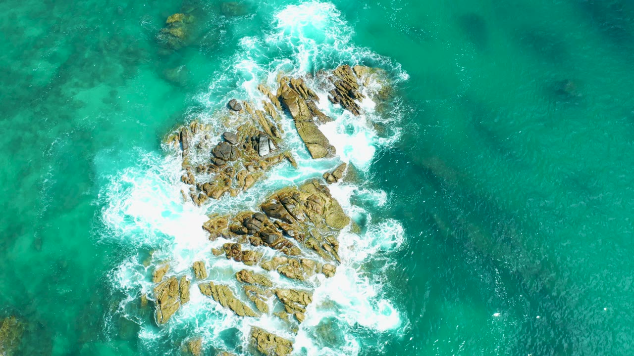 A rocky formation in green-blue water with foam curling around its left side.