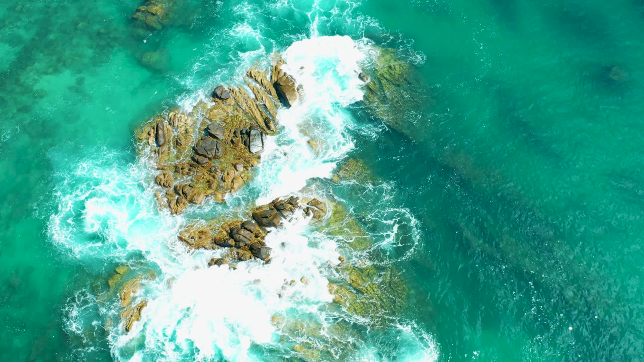 Aerial frame at Marquis Los Cabos, Mexico showing white wave foam spreading across the upper and lower edges of the rocky outcrop, with no visible people.