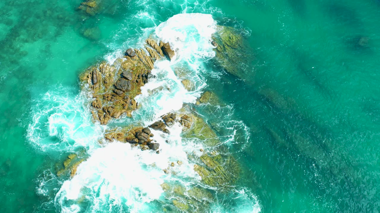 Aerial view at Marquis Los Cabos, Mexico with a foam swirl to the left of the rocks and breaking water across the top and lower rock faces, with no visible people.