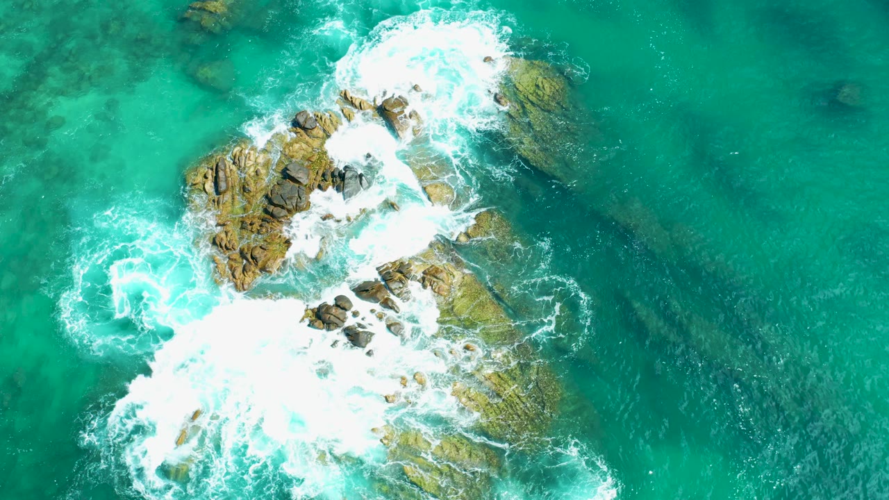 Aerial frame at Marquis Los Cabos, Mexico where the main rocks sit slightly left of center and heavy white foam washes across the lower section, with no visible people.