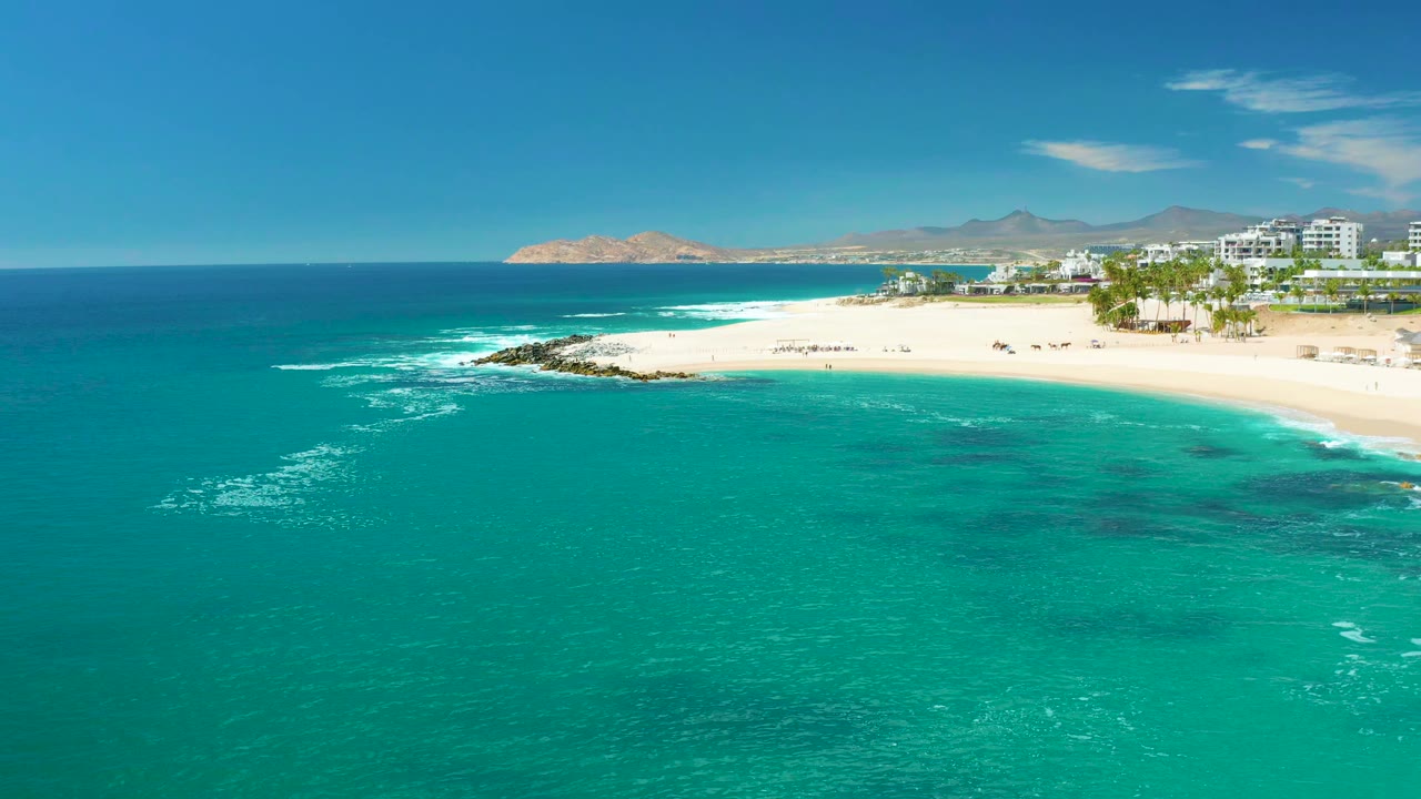 Aerial view at Marquis Los Cabos, Mexico showing turquoise water in the foreground, white surf near a sandy beach, rocks at the shoreline, coastal buildings to the right, distant mountains, and no visible people.