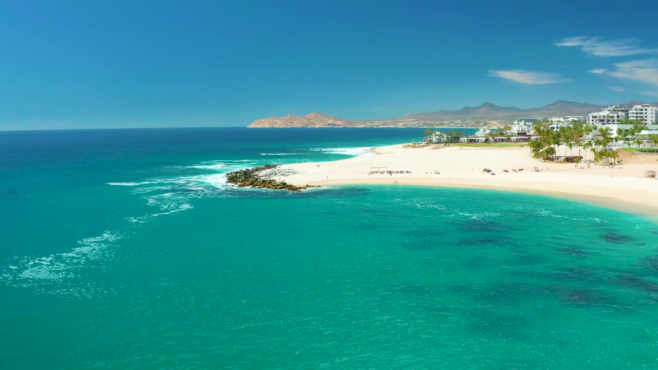 Aerial beach view with teal water, foamy surf, sand, rock jetty, buildings, palms, and distant hills.
