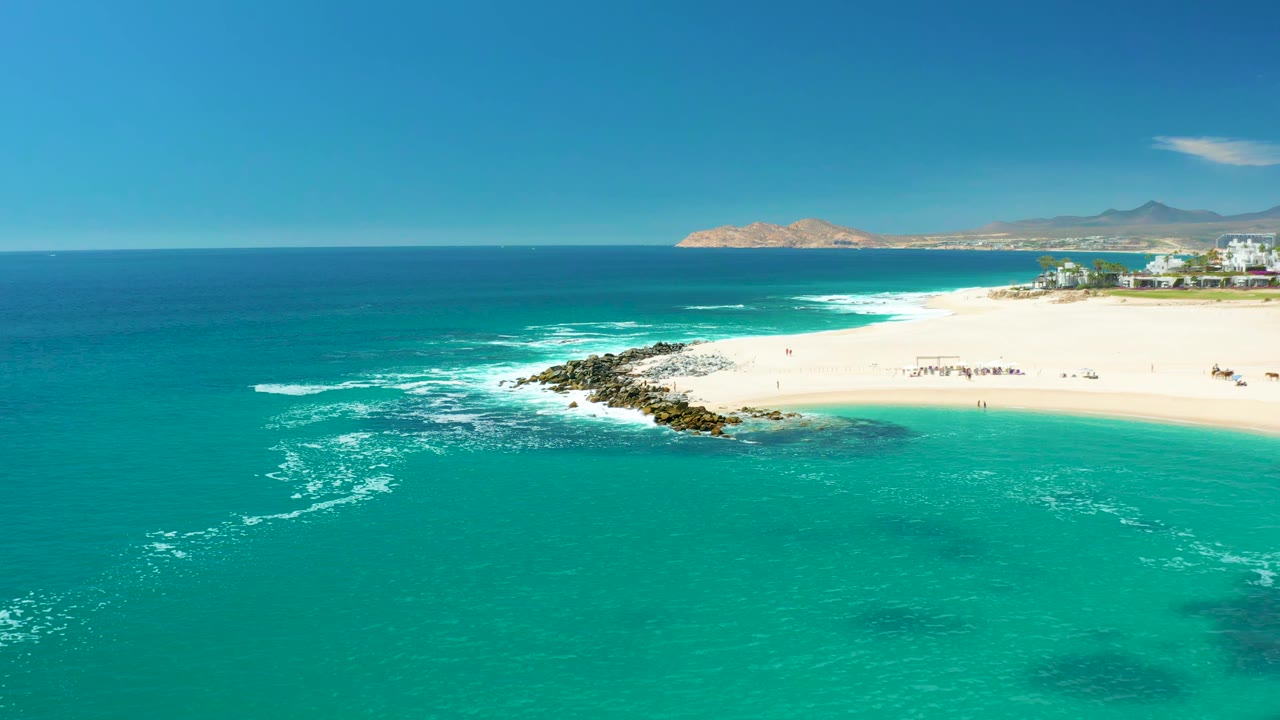 Turquoise water and white surf beside a sandy beach with people and distant buildings.