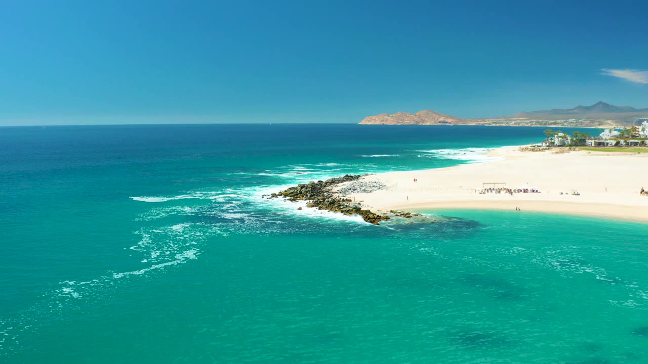 Wide drone-style frame over Marquis Los Cabos showing more open turquoise water in front, surf curling around rocks, a bright sandy beach with tiny beachgoers, and coastline buildings at the far right.